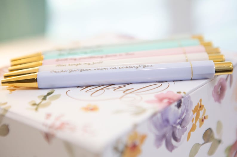 May include: A set of pastel colored pens with gold accents and inspirational quotes. The pens are arranged on a floral patterned box. The quote on the pen in focus reads "Praise God from whom all blessings flow."