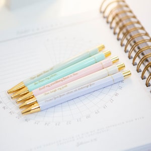 May include: Six pastel-colored pens with gold accents and inspirational quotes printed on them. The pens are arranged on a white page with a grid pattern.