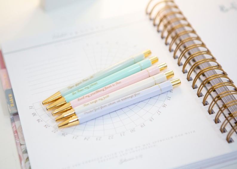 May include: Six pastel-colored pens with gold accents and inspirational quotes printed on them. The pens are arranged on a white page with a grid pattern.