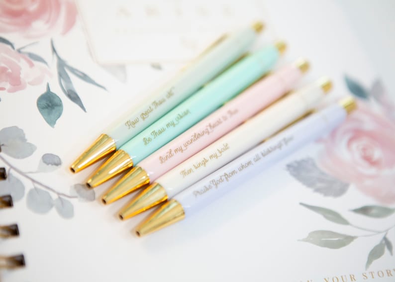 May include: Set of six pastel-colored pens with gold accents. Each pen has a different inspirational quote engraved on it. The quotes are: "How Great Thou Art", "Be Thou My Vision", "Bind My Wandering Heart To Thee", "Then Sings My Soul", "Praise God From Whom All Blessings Flow", and "Your Story".