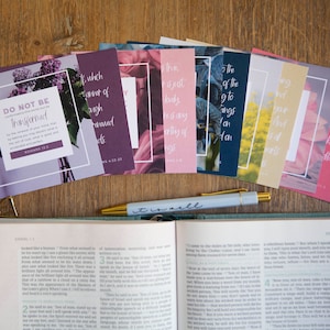 May include: A set of eight inspirational cards with floral designs and bible verses. The cards feature a variety of colors, including purple, pink, blue, and yellow. The cards are arranged in a row on a wooden surface, with a pen and an open bible in the background.
