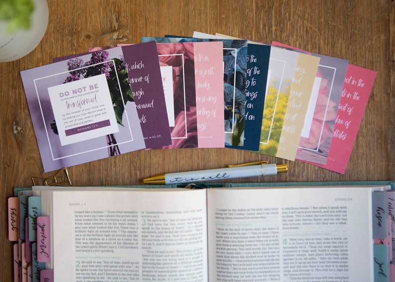May include: A set of eight inspirational cards with floral designs and bible verses. The cards feature a variety of colors, including purple, pink, blue, and yellow. The cards are arranged in a row on a wooden surface, with a pen and an open bible in the background.