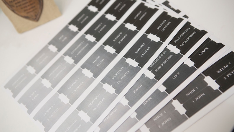 May include: A set of gray and black labels with the names of books of the Bible printed on them. The labels are arranged in a fan shape.