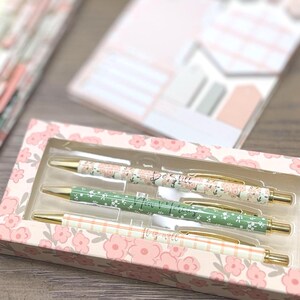 May include: A set of three retractable ballpoint pens housed in a rectangular cardboard box with a floral pattern. The pens feature gold-toned clips and tips, with barrels displaying distinct designs: one floral, one green with white flowers, and one plaid.