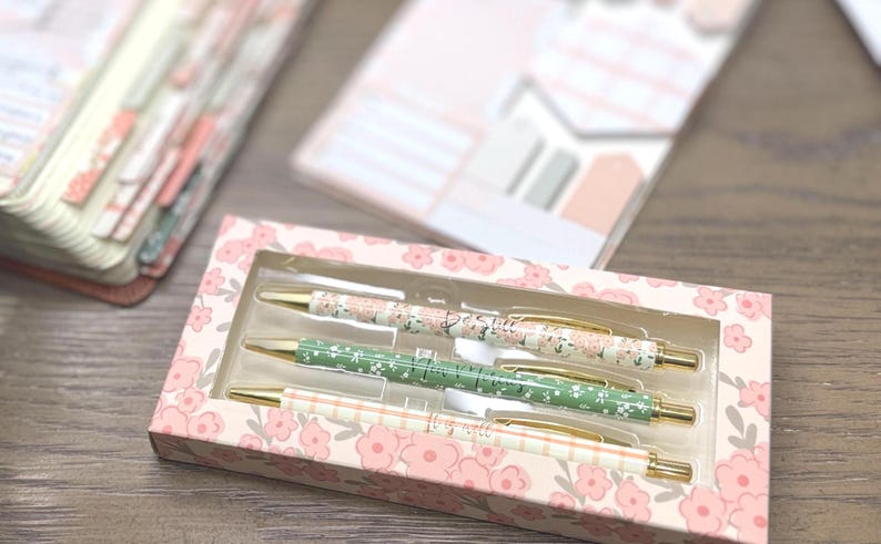 May include: A set of three retractable ballpoint pens housed in a rectangular cardboard box with a floral pattern. The pens feature gold-toned clips and tips, with barrels displaying distinct designs: one floral, one green with white flowers, and one plaid.
