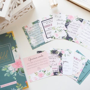 May include: A set of floral-patterned cards with inspirational quotes and bible verses. The cards are printed on white cardstock with black and pink lettering. The cards are arranged on a white surface with a teal blue journal with the word "Behold" on the cover.