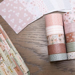 Pode incluir: Uma variedade de rolos de fita washi decorativa em v&aacute;rios designs florais e estampados, incluindo rosa, castanho e verde. Tamb&eacute;m s&atilde;o vis&iacute;veis folhas de papel coordenadas e um caderno com abas rotuladas, ideal para artesanato.