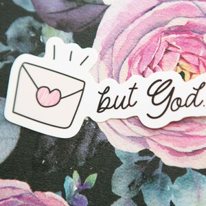 May include: White sticker with a pink envelope with a heart and the text "but God." The sticker is on a black background with pink flowers.