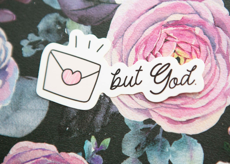 May include: White sticker with a pink envelope with a heart and the text "but God." The sticker is on a black background with pink flowers.