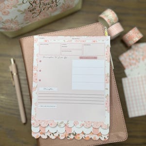 May include: A pink planner with floral accents, a pen, and decorative tape rolls. The planner has a floral design and the text "Principles to Live By". The background includes a floral makeup bag.