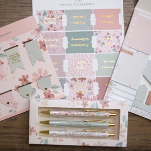 Peut inclure: Un ensemble de fournitures de papeterie &agrave; th&egrave;me floral rose et vert. L'ensemble comprend un paquet de signets, un paquet d'autocollants, un paquet de notes adh&eacute;sives et trois stylos. Les stylos ont un motif floral et le texte "Our Hope is Built on Nothing Less" imprim&eacute; dessus.