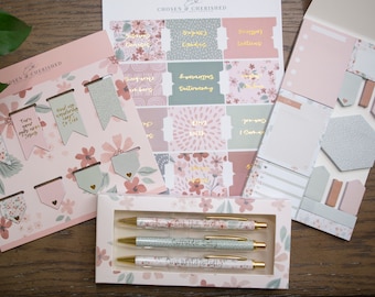 May include: A set of pink and green floral themed stationery supplies. The set includes a pack of bookmarks, a pack of stickers, a pack of sticky notes, and three pens. The pens have a floral design and the text "Our Hope is Built on Nothing Less" printed on them.