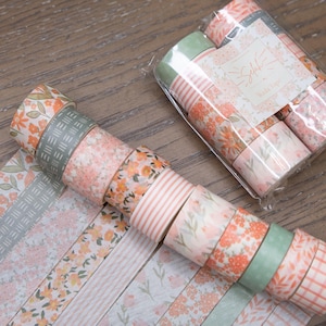 May include: A collection of decorative washi tapes in various floral and striped patterns. The tapes feature shades of peach, pink, and green, with some rolls still in their original packaging. Perfect for scrapbooking and crafting.