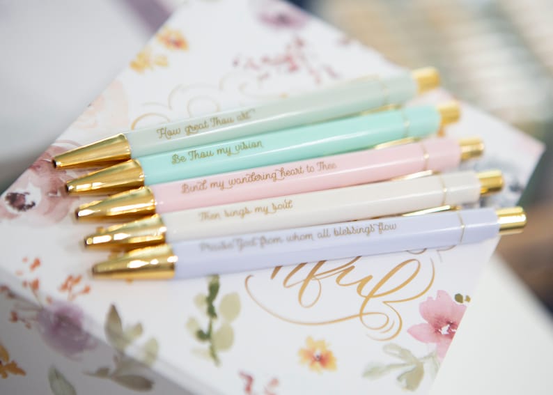 May include: A set of six pastel-colored pens with gold accents and inspirational quotes in gold lettering. The quotes are: "How great Thou art", "Be Thou my vision", "Bind my wandering heart to Thee", "Then sing my soul", "Praise God from whom all blessings flow".