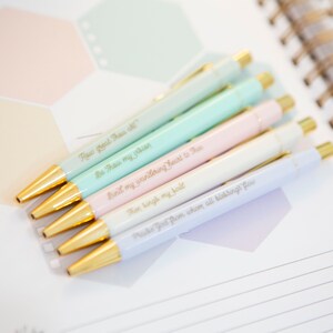 May include: Six pastel-colored pens with gold accents and inspirational quotes in gold lettering. The quotes are: "How Great Thou Art", "Be Thou my vision", "Bind my wandering heart to Thee", "Then bring my soul", "Praise God from whom all blessings flow".