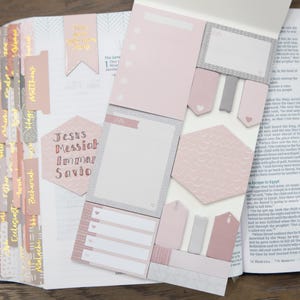 May include: A pink and gray Bible with a variety of pink and gray sticky notes. The sticky notes have different shapes and sizes. The Bible has a gold tab index with the names of the books of the Bible written in gold. The sticky notes have the words "Jesus Messiah Lord Savior" written on them.