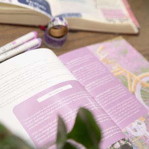 May include: An open book with pink pages and text, a gold paperclip, and two white pens with pink accents. A roll of decorative tape and a journal are also visible. The theme is stationery and journaling.