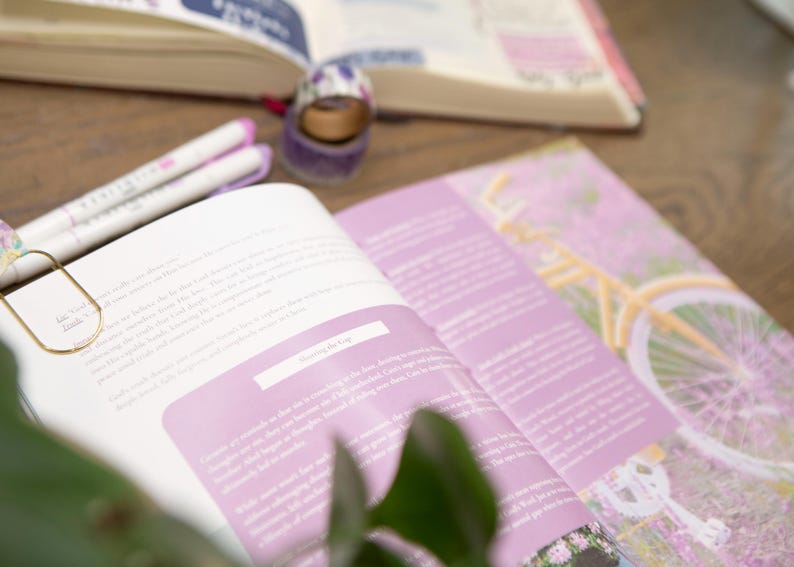 May include: An open book with pink pages and text, a gold paperclip, and two white pens with pink accents. A roll of decorative tape and a journal are also visible. The theme is stationery and journaling.