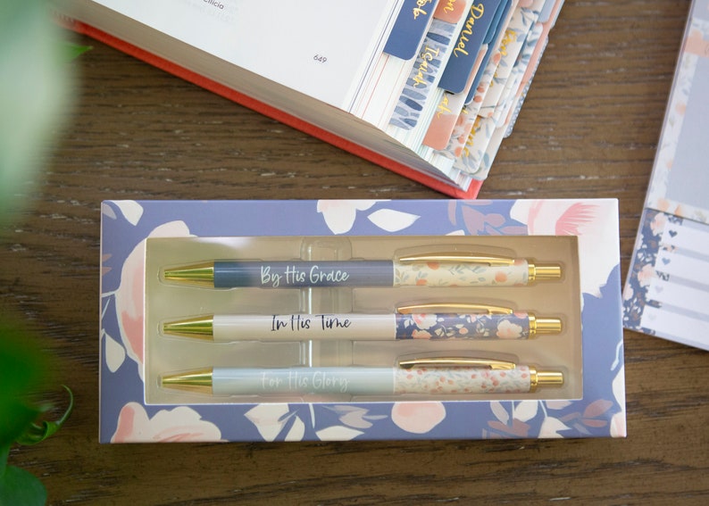 May include: A set of three blue and white pens with gold accents in a floral patterned box. The pens have the following text: "By His Grace", "In His Time", and "For His Glory".