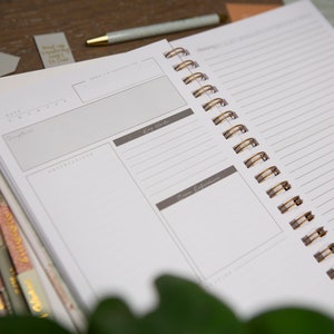 May include: An open spiral bound notebook with a white cover and gold wire binding. The notebook has a section for notes, key words, and source references. The notebook is lying on a wooden table.