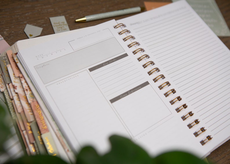 May include: An open spiral bound notebook with a white cover and gold wire binding. The notebook has a section for notes, key words, and source references. The notebook is lying on a wooden table.
