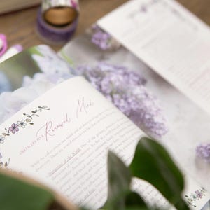 May include: An open book with the title "Renewed Mind" in cursive script, surrounded by floral illustrations. The pages display text and images of purple flowers. A roll of purple washi tape and pens are in the background.