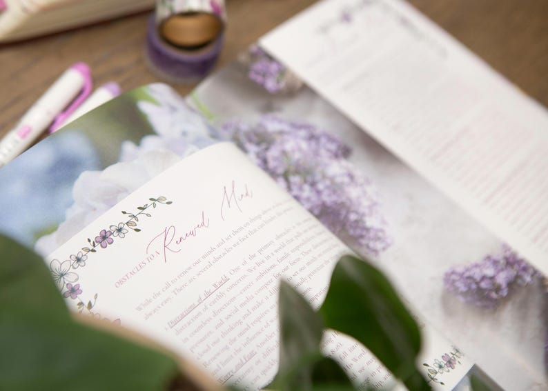 May include: An open book with the title "Renewed Mind" in cursive script, surrounded by floral illustrations. The pages display text and images of purple flowers. A roll of purple washi tape and pens are in the background.