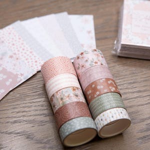 Pode incluir: Uma cole&ccedil;&atilde;o de artigos de papelaria com tema floral. A imagem apresenta rolos de fita adesiva decorativa washi em v&aacute;rios padr&otilde;es e cores, juntamente com uma pilha de pequenos blocos de notas e folhas de papel estampadas. A paleta de cores inclui rosas, castanhos e verdes.