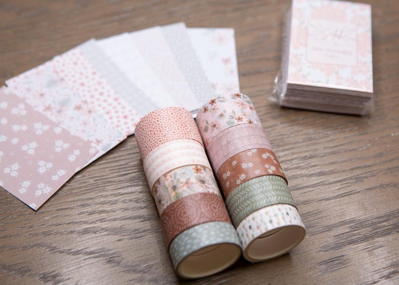 Pode incluir: Uma cole&ccedil;&atilde;o de artigos de papelaria com tema floral. A imagem apresenta rolos de fita adesiva decorativa washi em v&aacute;rios padr&otilde;es e cores, juntamente com uma pilha de pequenos blocos de notas e folhas de papel estampadas. A paleta de cores inclui rosas, castanhos e verdes.