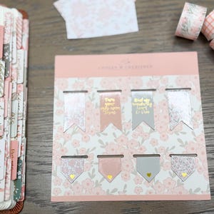 May include: A set of eight magnetic bookmarks on a floral backing card, featuring pink and gray colors with gold foil text and heart accents. The bookmarks are arranged in two rows, with some displaying text and others featuring floral or geometric patterns.