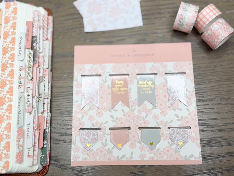 May include: A set of eight magnetic bookmarks on a floral backing card, featuring pink and gray colors with gold foil text and heart accents. The bookmarks are arranged in two rows, with some displaying text and others featuring floral or geometric patterns.