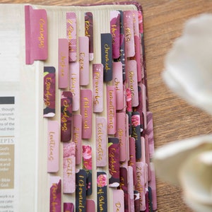 May include: A close-up of a Bible with pink and burgundy floral patterned tabs. The tabs are labeled with the names of books of the Bible, including Genesis, Exodus, Leviticus, Numbers, Deuteronomy, Joshua, Judges, Ruth, 1 Samuel, 2 Samuel, 1 Kings, 2 Kings, 1 Chronicles, 2 Chronicles, Ezra, Nehemiah, Esther, Job, Psalms, Proverbs, Ecclesiastes, Song of Solomon, Isaiah, Jeremiah, Lamentations, Ezekiel, Daniel, Hosea, Joel, Amos, Obadiah, Jonah, Micah, Nahum, Habakkuk, Zephaniah, Haggai, Zechariah, Malachi, Matthew, Mark, Luke, John, Acts, Romans, 1 Corinthians, 2 Corinthians, Galatians, Ephesians, Philippians, Colossians, 1 Thessalonians, 2 Thessalonians, 1 Timothy, 2 Timothy, Titus, Philemon, Hebrews, James, 1 Peter, 2 Peter, 1 John, 2 John, 3 John, Jude, and Revelation.