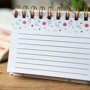 May include: A white spiral bound notebook with a floral design on the cover. The notebook has lined pages and is perfect for taking notes or writing down ideas.