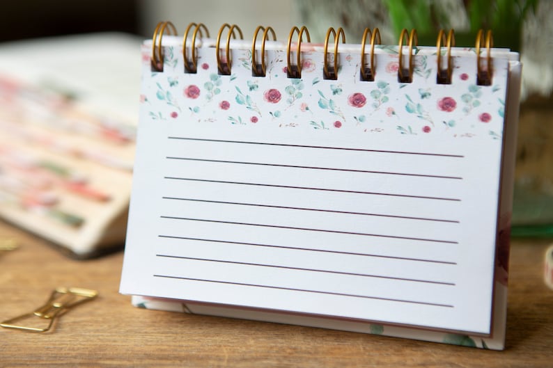 May include: A white spiral bound notebook with a floral design on the cover. The notebook has lined pages and is perfect for taking notes or writing down ideas.