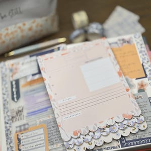 May include: A journal with a pink and floral design, open to a page with a pink paper insert. The insert has the words "Strengths" and "Weaknesses" printed on it. A small pouch with the words "be still" is in the background.