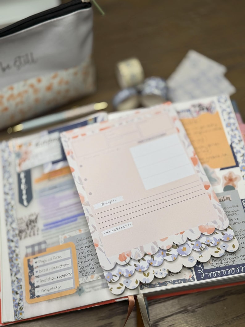 May include: A journal with a pink and floral design, open to a page with a pink paper insert. The insert has the words "Strengths" and "Weaknesses" printed on it. A small pouch with the words "be still" is in the background.