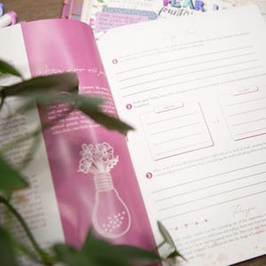 May include: Open journal with pink accents and lined pages for notes and reflections. The left page features a lightbulb design with flowers. Pens are visible in the background. The journal is open to a page with prompts for writing.