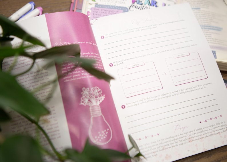 May include: Open journal with pink accents and lined pages for notes and reflections. The left page features a lightbulb design with flowers. Pens are visible in the background. The journal is open to a page with prompts for writing.