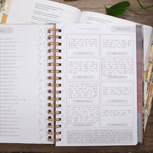 Op de afbeelding: Een witte en grijze planner met een gouden spiraalbinding. De planner heeft een leesschema met een lijst van bijbelverzen en een sectie met bijbelverzen en instructies. De planner is open op een pagina met de woorden "Fearless" en "The Word" geschreven in cursief.