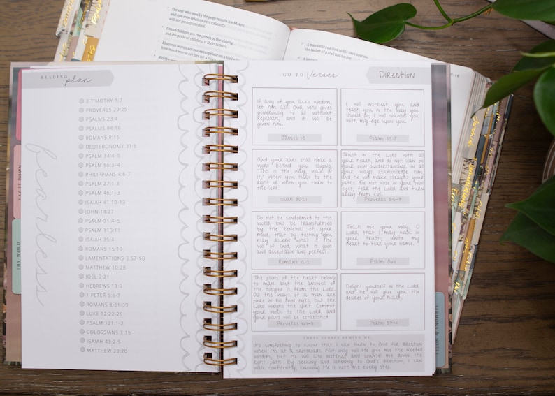 Op de afbeelding: Een witte en grijze planner met een gouden spiraalbinding. De planner heeft een leesschema met een lijst van bijbelverzen en een sectie met bijbelverzen en instructies. De planner is open op een pagina met de woorden "Fearless" en "The Word" geschreven in cursief.