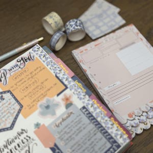May include: A planner with floral accents, washi tape rolls, and a pen. The planner pages have handwritten notes and stickers. The washi tape rolls have floral and plaid patterns. The pen has a floral design.