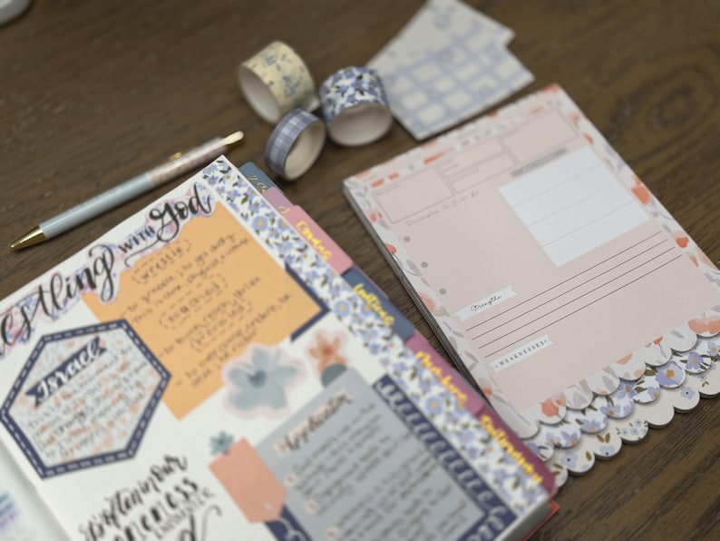 May include: A planner with floral accents, washi tape rolls, and a pen. The planner pages have handwritten notes and stickers. The washi tape rolls have floral and plaid patterns. The pen has a floral design.