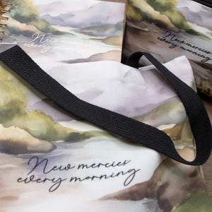 Puede incluir: Una bolsa de tela blanca con correas negras y un dise&ntilde;o de acuarela de monta&ntilde;as y un r&iacute;o. La bolsa tiene el texto "New mercies every morning" impreso en ella.