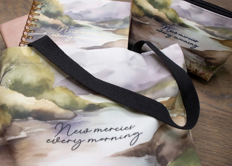 Puede incluir: Una bolsa de tela blanca con correas negras y un dise&ntilde;o de acuarela de monta&ntilde;as y un r&iacute;o. La bolsa tiene el texto "New mercies every morning" impreso en ella.