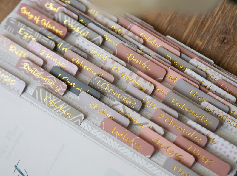 May include: A set of pink and gray dividers with gold lettering for a Bible. The dividers are labeled with the names of the books of the Bible, including Genesis, Exodus, Leviticus, Numbers, Deuteronomy, Joshua, Judges, Ruth, 1 Samuel, 2 Samuel, 1 Kings, 2 Kings, 1 Chronicles, 2 Chronicles, Ezra, Nehemiah, Esther, Job, Psalms, Proverbs, Ecclesiastes, Song of Solomon, Isaiah, Jeremiah, Lamentations, Ezekiel, Daniel, Hosea, Joel, Amos, Obadiah, Jonah, Micah, Nahum, Habakkuk, Zephaniah, Haggai, Zechariah, Malachi.