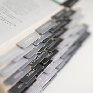 May include: A close-up of a Bible with black and gray tabs marking the different books of the Bible. The tabs are labeled with the names of the books, including Romans, Daniel, Song of Solomon, Chronicles, Numbers, and Isaiah.