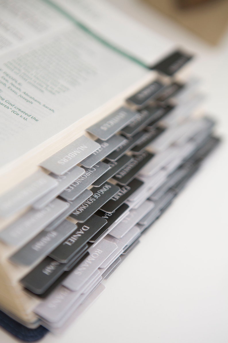 May include: A close-up of a Bible with black and gray tabs marking the different books of the Bible. The tabs are labeled with the names of the books, including Romans, Daniel, Song of Solomon, Chronicles, Numbers, and Isaiah.