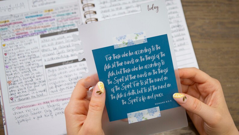 May include: A blue card with a white border and floral patterned tape. The card has a quote from Romans 8:6: "For those who live according to the flesh set their minds on the things of the flesh, but those who live according to the Spirit set their minds on the things of the Spirit. For to set the mind on the flesh is death, but to set the mind on the Spirit is life and peace."
