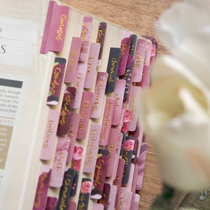 May include: A close-up of a pink and gold Bible with floral patterned tabs. The tabs are labeled with the names of books in the Bible, including Genesis, Exodus, Leviticus, Numbers, Deuteronomy, Joshua, Judges, Ruth, 1 Samuel, 2 Samuel, 1 Kings, 2 Kings, 1 Chronicles, 2 Chronicles, Ezra, Nehemiah, Esther, Job, Psalms, Proverbs, Ecclesiastes, Song of Solomon, Isaiah, Jeremiah, Lamentations, Ezekiel, Daniel, Hosea, Joel, Amos, Obadiah, Jonah, Micah, Nahum, Habakkuk, Zephaniah, Haggai, Zechariah, Malachi.