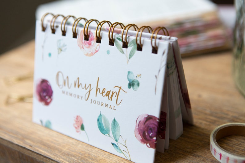 May include: A small white spiral bound journal with a floral design and gold lettering that reads "On my heart Memory Journal".
